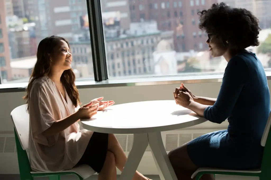 Two women talking across a desk in a bright office setting.